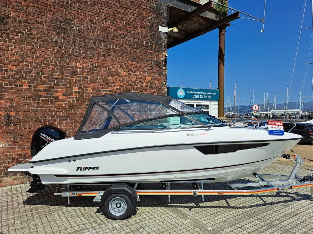 A white Flipper 650 DC boat for sale with a visible "FOR SALE" sign, parked near a brick wall and marina.